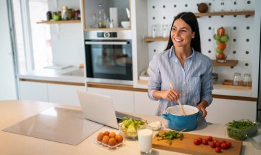 Bir kadın mutfakta, elinde bir bardak süt tutarken gülümsüyor ve dizüstü bilgisayarından tarifine bakıyor. Taze yapraklı sebzeler tezgahın üzerinde oturur.