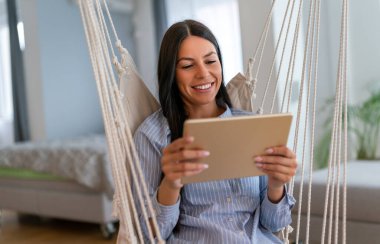 Sosyal medya zamanı. Tabletiyle temasta kalmak. Evdeki oturma odasında tablet tablet kullanan neşeli bir kadın..