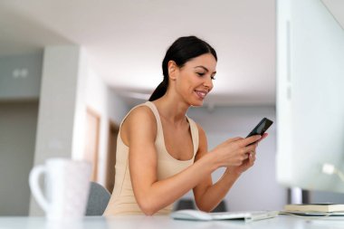 Genç ve güzel bir kadın sosyal medyayı kontrol etmek, çalışmak veya e-postaları okumak için bilgisayar kullanıyor. Dijital cihazlarla evden çalışan yaratıcı iş adamı.