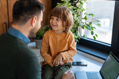 Mutlu aile fotoğrafı. Oğlu gülerken ve birlikte eğlenirken, genç yakışıklı adam laptopta evden çalışıyor.