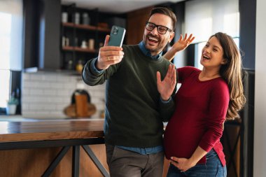 Kocası cep telefonuyla selfie çekerken evde gülümseyen hamile bir kadın. Genç çiftin fotoğraf çektiği romantik bir aile anı. Ebeveyn olmak için sabırsızlanan mutlu bir çift.