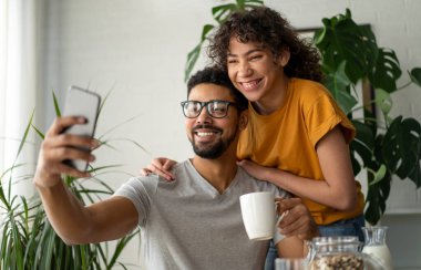 Neşeli siyah çift evde kahvaltı ederken gülümsüyor ve cep telefonuyla selfie çekiyor.