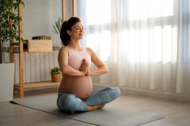 Rahatlamış hamile bir kadın evde egzersiz minderinde bacak bacak üstüne atarak otururken nefes egzersizi yapıyor..
