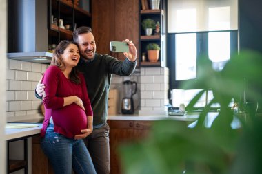 Sevimli bir adam hamile karısına sarılıyor, hamileliğinin son üç ayında selfie çekerek eğleniyor. Mutlu koca hamile kadını kucaklarken bebek bekliyor..
