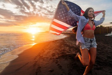 Güzel vatansever, hayat dolu genç bir kadın. Amerikan bayrağı uzanmış ellerinde. Yaz günbatımında ayakta duruyor.