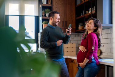 Hamilelik, teknoloji ve insan kavramı. Mutlu adam ve evde akıllı telefonları olan hamile karısı. Yeni mutlu aile.