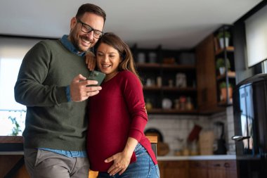 Hamilelik, teknoloji ve insan kavramı. Mutlu adam ve evde akıllı telefonları olan hamile karısı. Yeni mutlu aile.