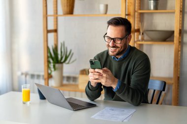 Elektronik bankacılık için akıllı telefon ve dizüstü bilgisayar kullanan, rezervasyon yapan, online alışveriş yapan ve evde vakit geçirirken ödeme yapan şık giyimli bir adam..