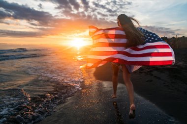 Güzel vatansever, hayat dolu genç bir kadın. Amerikan bayrağı uzanmış ellerinde. Yaz günbatımında ayakta duruyor.