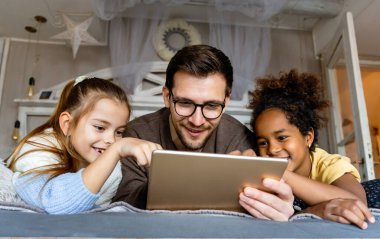 Evde dijital tabletle oynayan genç bekar baba ve gülümseyen kızlar. Dijital tablet kullanan neşeli ve heyecanlı kızları olan genç ebeveynler.