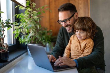 Mutlu aile fotoğrafı. Oğlu gülerken ve birlikte eğlenirken, genç yakışıklı adam laptopta evden çalışıyor.