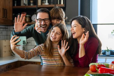 Mutlu ebeveynler ve iki çocuk birlikte akıllı telefon kullanıyorlar, gülümseyen anne ve baba ve küçük oğlu ve kızı telefon ekranına bakıyor, selfie çekiyor, eğleniyorlar.