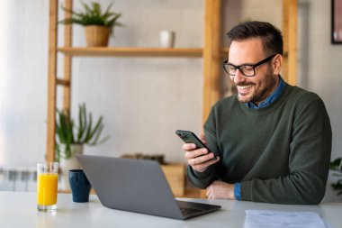Mutlu gülümseyen iş adamı günlük kıyafetler giyer ve ev ofisinde modern akıllı telefon kullanır. İnsanlar dijital cihazlar modern yaşam konsepti