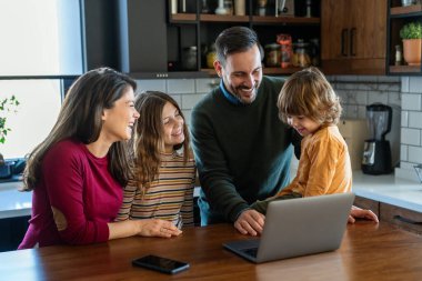 Mutfakta çocuklu mutlu bir aile birlikte modern dizüstü bilgisayar kullanarak eğleniyor, gülümseyen ebeveynler çocuklarla birlikte hafta sonunun keyfini çıkarıyor evde bilgisayar başında gülen videolar izliyorlar.