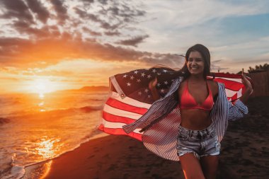 Güzel vatansever, hayat dolu genç bir kadın. Amerikan bayrağı uzanmış ellerinde. Yaz günbatımında ayakta duruyor.