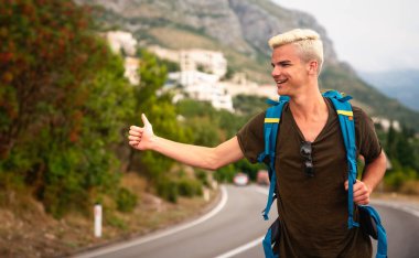 Yalnız seyahat etme kavramı. Güneşli bir yaz gününde yolda otostop çeken genç beyaz bir adam. Sırt çantalı hippi dağlarda arabayı durdurmaya çalışıyor.
