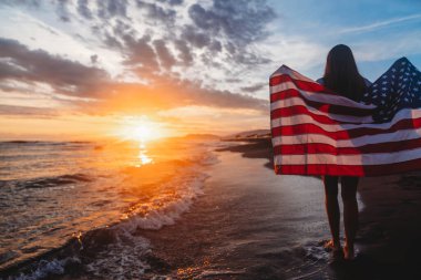Güzel vatansever, hayat dolu genç bir kadın. Amerikan bayrağı uzanmış ellerinde. Yaz günbatımında ayakta duruyor.