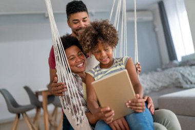 Happy african family having fun with device at home, black parents and child using digital tablet looking at screen together. clipart