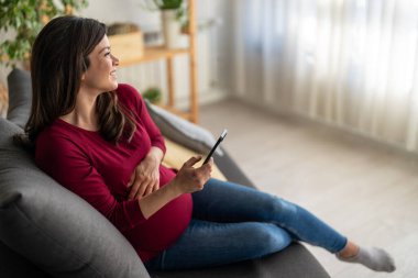 Hamile bir kadın online hamilelik öncesi ders için telefon uygulaması kullanarak yatakta dinleniyor. Mobil ve bebek göbeğine odaklan.