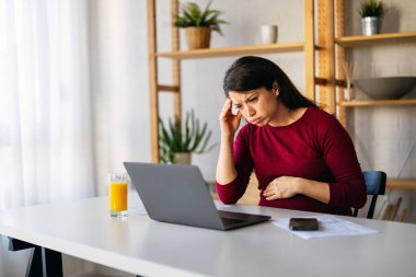 Güzel, beyaz, hamile bir kadın karın ağrısı çekiyor. Evde dizüstü bilgisayarla çalışırken rahatlamaya çalışıyor. Son üç aylık dönemde karın ağrısından muzdarip olan bir bayanın krampları var.