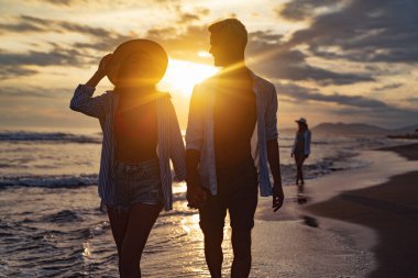 Genç ve güzel bir çift gün batımında deniz kıyısında yürüyor. Balayı. Romantik gezi..