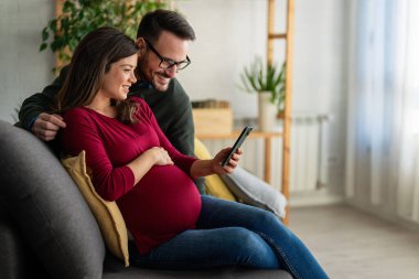 Mutlu çift evde sarılıp arkadaşlarıyla, aile üyeleriyle video görüşmesi yapıyor. Neşeli hamile kadın ve kocası iyi ruh hali içinde ailelerini selamlıyor, uygulamayla internet üzerinden iletişim kuruyor.
