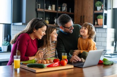 Mutfakta çocuklu mutlu bir aile birlikte modern dizüstü bilgisayar kullanarak eğleniyor, gülümseyen ebeveynler çocuklarla birlikte hafta sonunun keyfini çıkarıyor evde bilgisayar başında gülen videolar izliyorlar.