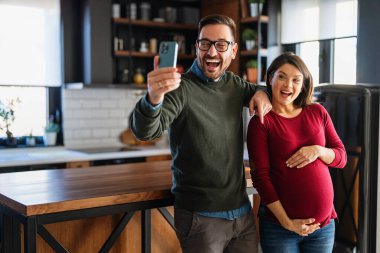 Mutlu çift evde sarılıp arkadaşlarıyla, aile üyeleriyle video görüşmesi yapıyor. Neşeli hamile kadın ve kocası iyi ruh hali içinde ailelerini selamlıyor, uygulamayla internet üzerinden iletişim kuruyor.
