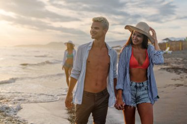 Kumsalda birlikte yürüyen tatilci çift birbirine sarılıyor. Mutlu ırklar arası genç çift yaz tatilinde balayının tadını çıkarıyor.