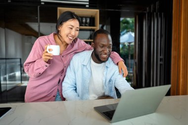 Çevrimiçi alışveriş veya ödeme için dijital dizüstü bilgisayar kullanan mutlu çift. Kadın kocasına dijital cihazdan bir şey alıyor. Orta Doğulu, Afro-Amerikalı çift dinleniyor..