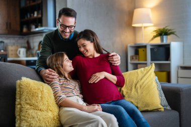 Sevgi dolu gülümseyen genç baba ve hamile anne portresi kanepeye oturmuş, neşeli çocukla eğleniyor, bebek bekliyor, samimi aile ilişkileri, çocuk bakımı konsepti.
