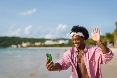 Mutlu Afrikalı genç Amerikalı adam tropikal bir plajda akıllı telefonuyla konuşuyor, tatilin, uzaktan çalışmanın, sosyal medyanın nişanının ve kaygısız dijital yaşam tarzının tadını çıkarıyor.