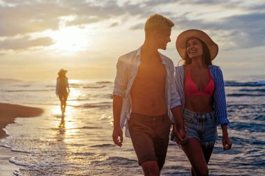 Genç ve güzel bir çift gün batımında deniz kıyısında yürüyor. Balayı. Romantik gezi..