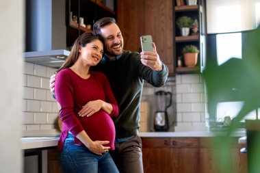 Hamilelik, teknoloji ve insan kavramı. Mutlu adam ve evde akıllı telefonları olan hamile karısı. Yeni mutlu aile.