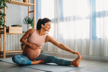 Aktif hamile kadın yoga yapıyor, kaslarını mindere geriyor, hamilelik öncesi egzersizleri yapıyor ve vücudu doğuma hazırlıyor.