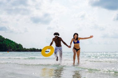 Mutlu ırklar arası bir çift, eğlenceli bir plaj gününün tadını çıkarıyor, okyanus dalgalarında su sıçratıyorlar, el ele tutuşuyorlar, ve sarı yüzme yüzüğüyle güneşli gökyüzünün altında gülümsüyorlar.