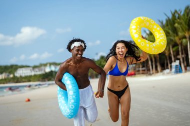 Irklar arası bir çift sahilde eğleniyor, okyanus dalgalarında koşuyor, gülümsüyor ve güneşli bir yaz gününde tropik bir tatilin tadını çıkarıyor.