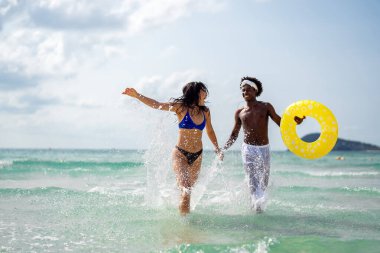 Irklar arası bir çift sahilde eğleniyor, okyanus dalgalarında koşuyor, gülümsüyor ve güneşli bir yaz gününde tropik bir tatilin tadını çıkarıyor.
