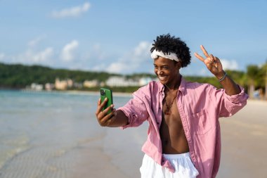 Mutlu Afrikalı genç Amerikalı adam tropikal bir plajda akıllı telefonuyla konuşuyor, tatilin, uzaktan çalışmanın, sosyal medyanın nişanının ve kaygısız dijital yaşam tarzının tadını çıkarıyor.