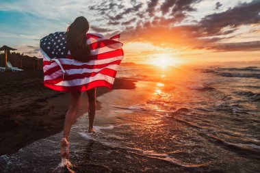 Gülümseyen ve sahilde koşan mutlu kadın. Amerikan bayrağı taşıyan bir kadının portresi. 4 Temmuz bağımsızlık günü. ABD 'de tatil. ABD sembolü. Gün batımı