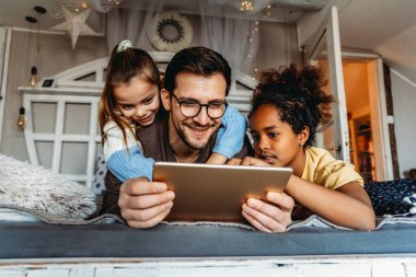 Bekar baba, evdeki dijital tabletle oturma odasındaki yatakta etnik çeşitlilikteki çocuklarıyla oynuyor.