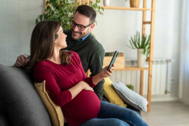 Hamilelik, teknoloji ve insan kavramı. Mutlu adam ve evde akıllı telefonları olan hamile karısı. Yeni mutlu aile.