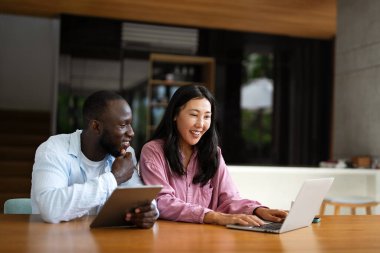 Çok uluslu çift sabah rutininin keyfini çıkarıyorlar, gülümsüyorlar ve rahat ve modern bir ev ortamında dizüstü bilgisayar ve tablet kullanıyorlar.