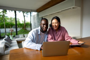 Çevrimiçi alışveriş veya ödeme için dijital dizüstü bilgisayar kullanan mutlu çift. Kadın kocasına dijital cihazdan bir şey alıyor. Orta Doğulu, Afro-Amerikalı çift dinleniyor..