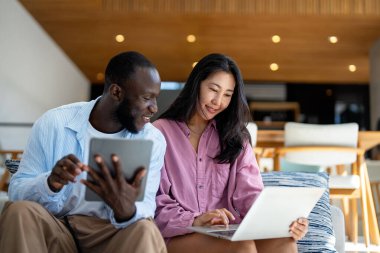 Çok uluslu çift sabah rutininin keyfini çıkarıyorlar, gülümsüyorlar ve rahat ve modern bir ev ortamında dizüstü bilgisayar ve tablet kullanıyorlar.