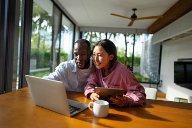 Çok uluslu çift sabah rutininin keyfini çıkarıyorlar, gülümsüyorlar ve rahat ve modern bir ev ortamında dizüstü bilgisayar ve tablet kullanıyorlar.