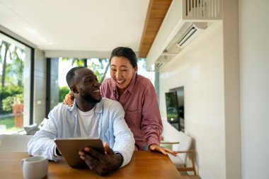 Genç ve çok ırklı çift evde tablet kullanırken eğleniyor. Tablet yazarken kucaklaşan genç insanları sevmek. Mutlu çift gelecek planlarını konuşuyor.