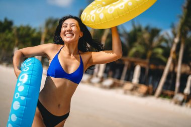 Heyecanlı genç kadın tropikal plaj tatilinin tadını çıkarıyor, elinde renkli şişirilebilir yüzükler tutuyor ve eğlenceli bir yaz kaçamağını kutluyor.