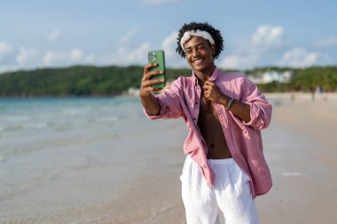 Mutlu Afrikalı genç Amerikalı adam tropikal bir plajda akıllı telefonuyla konuşuyor, tatilin, uzaktan çalışmanın, sosyal medyanın nişanının ve kaygısız dijital yaşam tarzının tadını çıkarıyor.