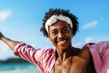 Genç siyahi adam güneşli bir plaj gününün tadını çıkarıyor, okyanus dalgalarında koşuyor, gülümsüyor ve berrak su ve mavi gökyüzü ile tropikal bir tatilde özgür hissediyor.